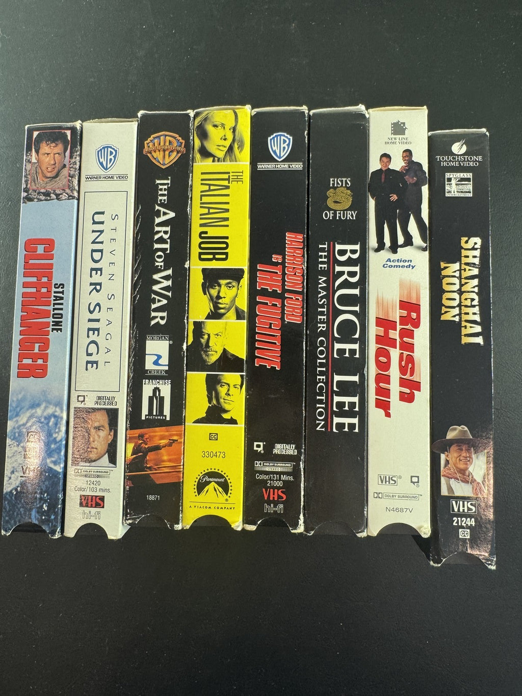 Collection of 8 vintage action movie VHS tapes including Rush Hour, Cliffhanger, and The Fugitive.