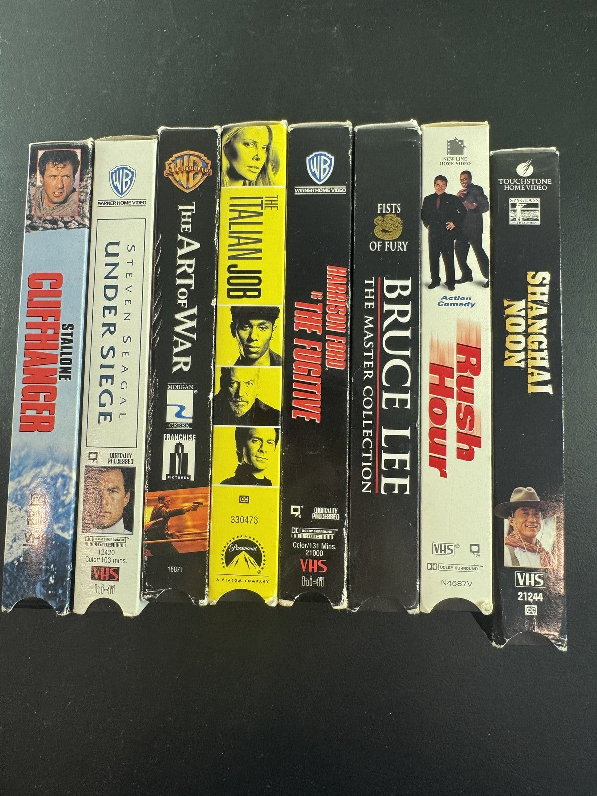 Collection of 8 vintage action movie VHS tapes including Rush Hour, Cliffhanger, and The Fugitive.