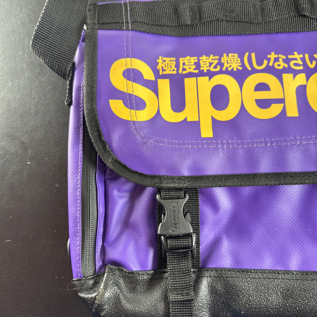Purple and yellow Superdry tarp messenger bag with bold logo spellout and padded shoulder strap.
