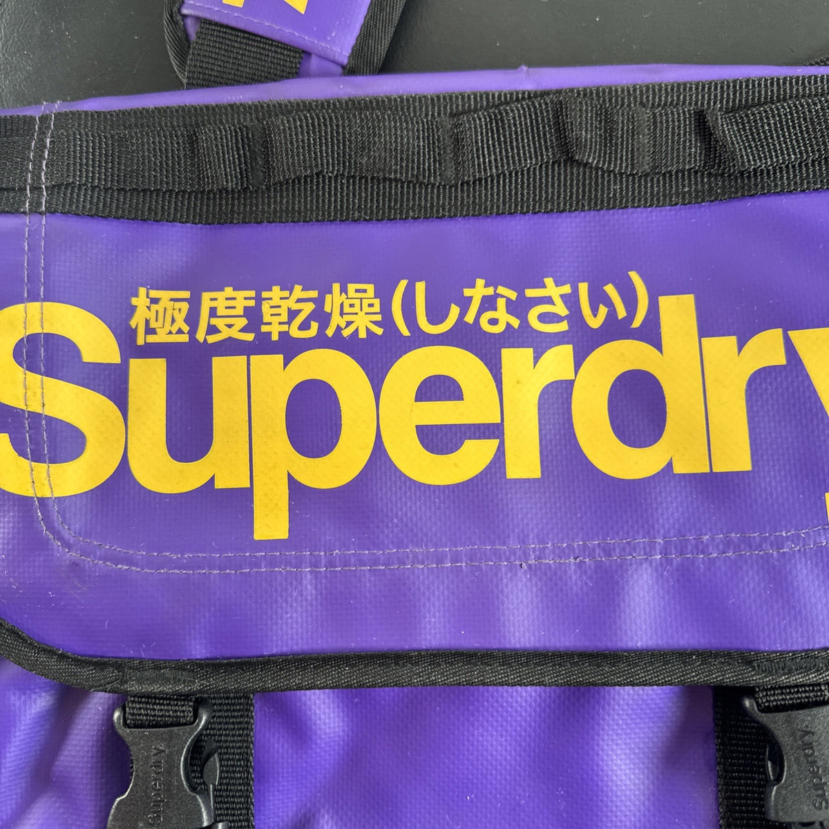 Purple and yellow Superdry tarp messenger bag with bold logo spellout and padded shoulder strap.
