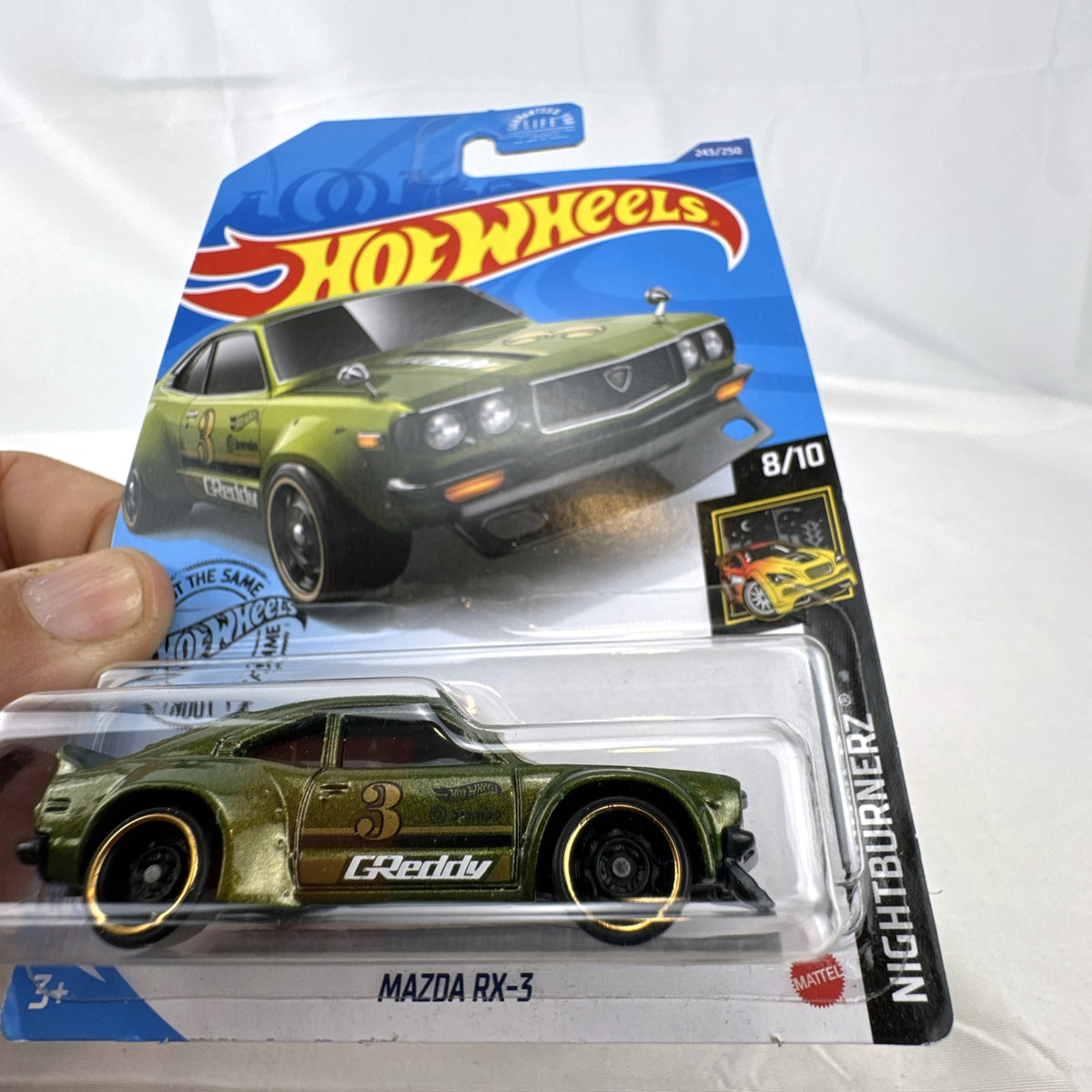 Hot Wheels 1:64 scale Mazda RX-3 diecast car in metallic olive green with GReddy graphics on blister card