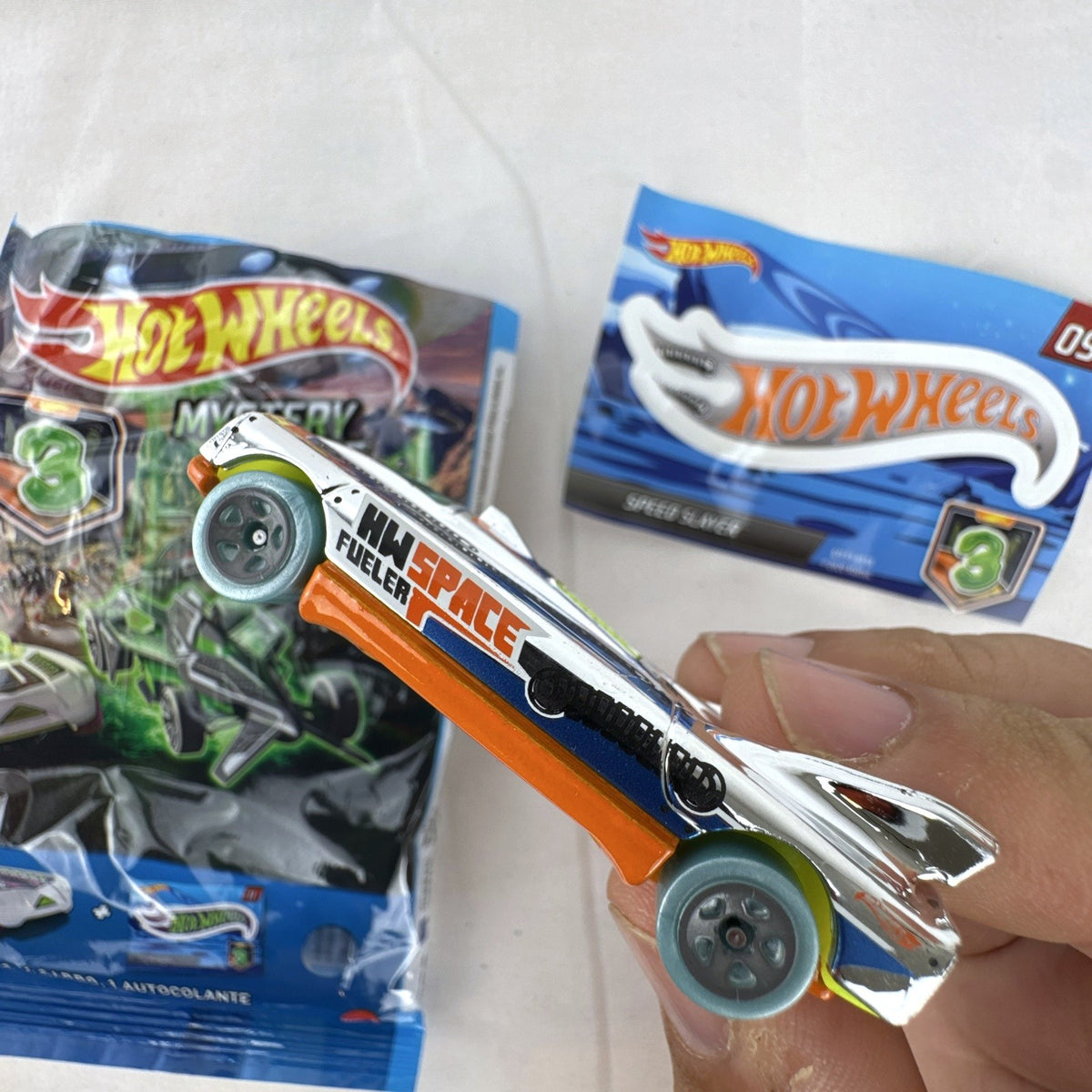 Hot Wheels Speed Slayer 1:64 scale diecast car in chrome with orange base, lime green canopy, foil bag, and sticker.