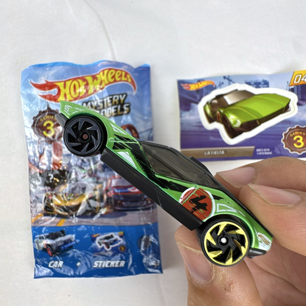 Metallic green Hot Wheels La Fasta 1:64 diecast car from Mystery Models Series 3 with original sticker card.