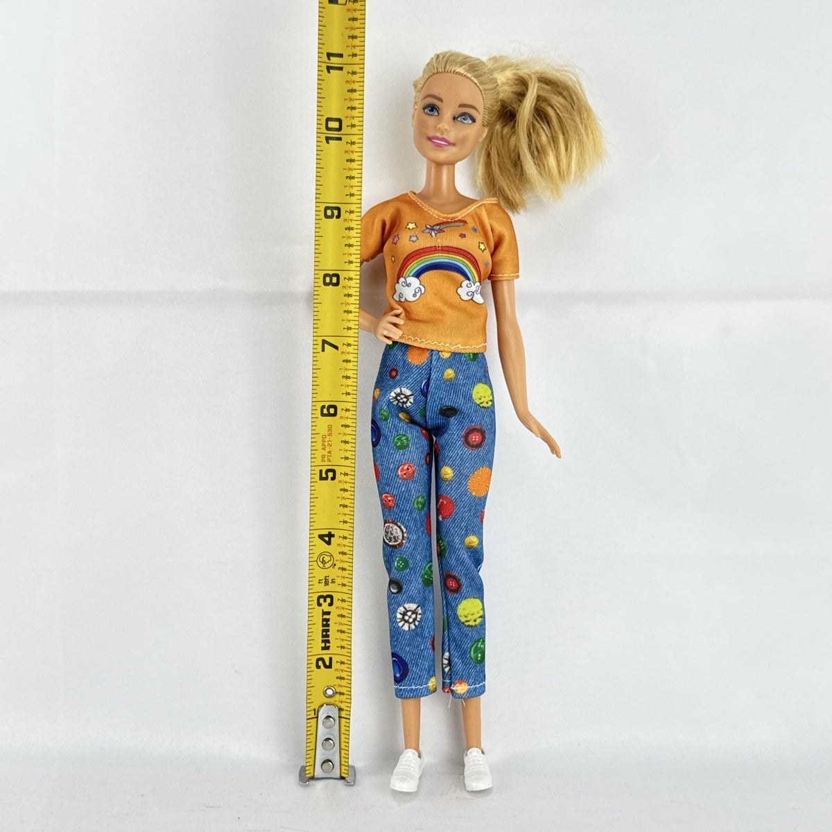 Mattel blonde Barbie doll wearing an orange rainbow shirt, button print pants, and white sneakers.