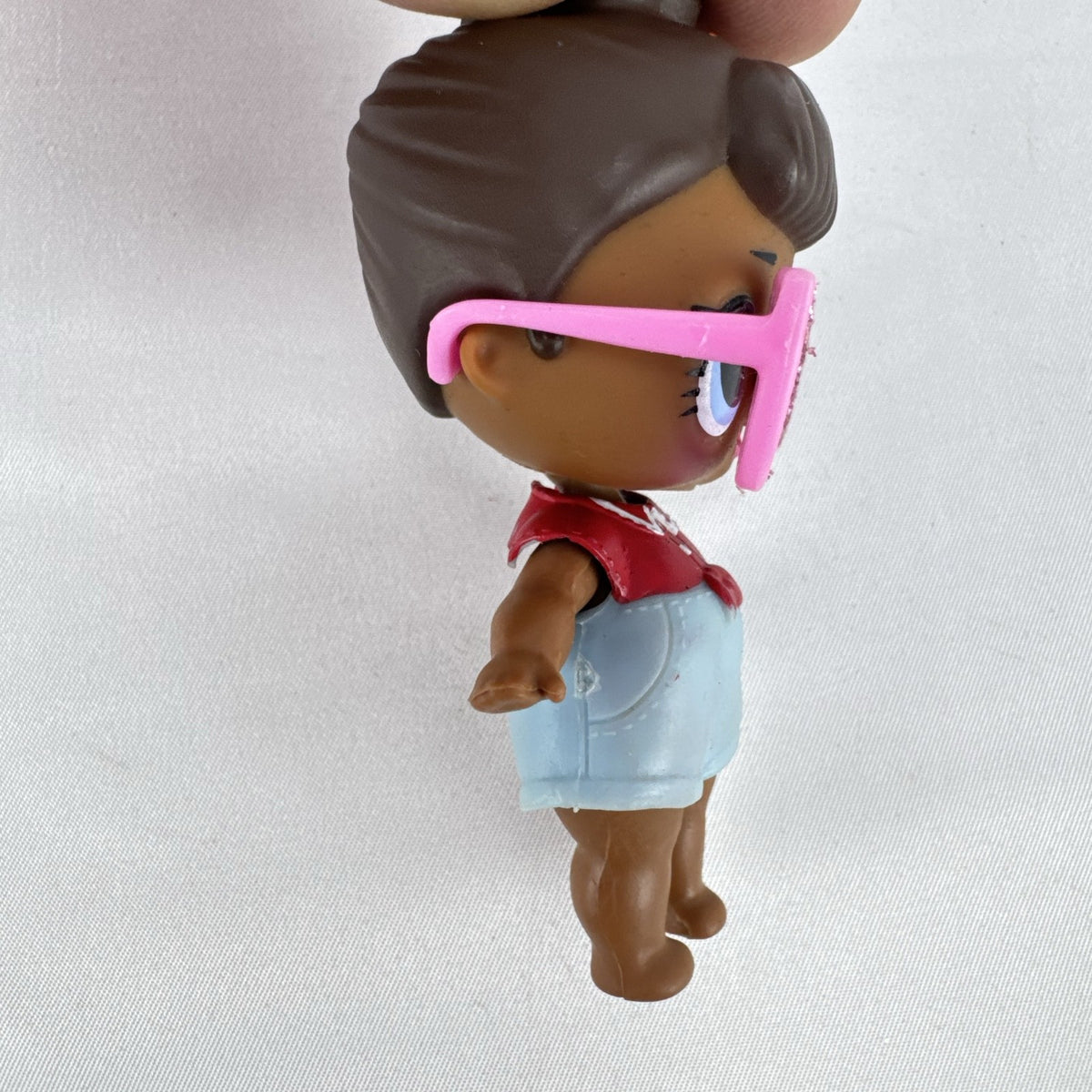 MGA L.O.L. Surprise! 3-inch doll with brown hair bun, pink glitter cat-eye glasses, and red tie-front outfit