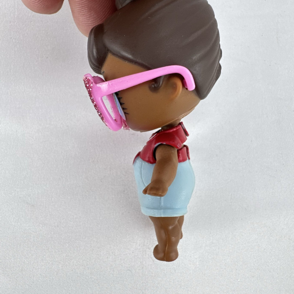 MGA L.O.L. Surprise! 3-inch doll with brown hair bun, pink glitter cat-eye glasses, and red tie-front outfit